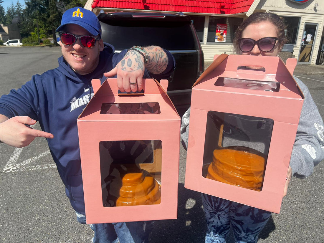 Two smiling people outdoors holding large pink display boxes with oversized caramel props inside.